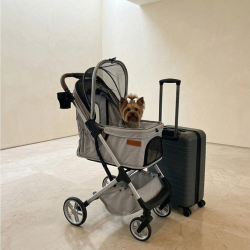A small dog sits inside a gray pet stroller with a mesh canopy, positioned next to a dark gray rolling suitcase in a minimalist beige room.