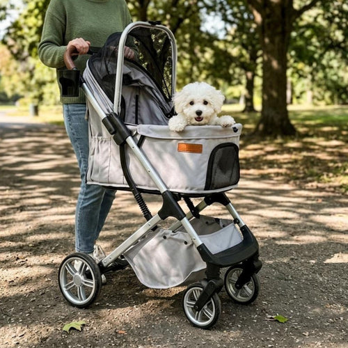 A small white dog sits in a pet stroller being pushed outdoors on a park path. The stroller has a light gray fabric body with black mesh panels and a silver metal frame.